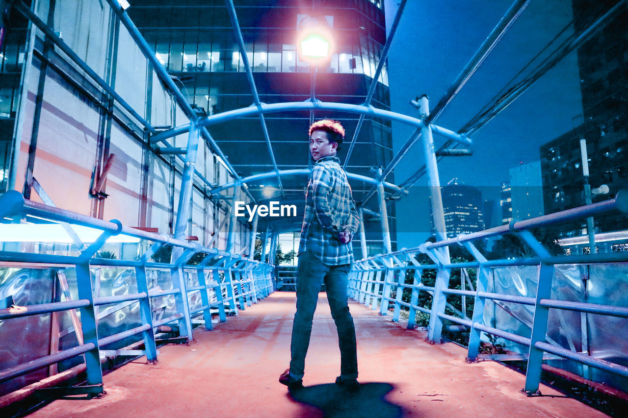 Full length portrait of man standing on footbridge in city at night