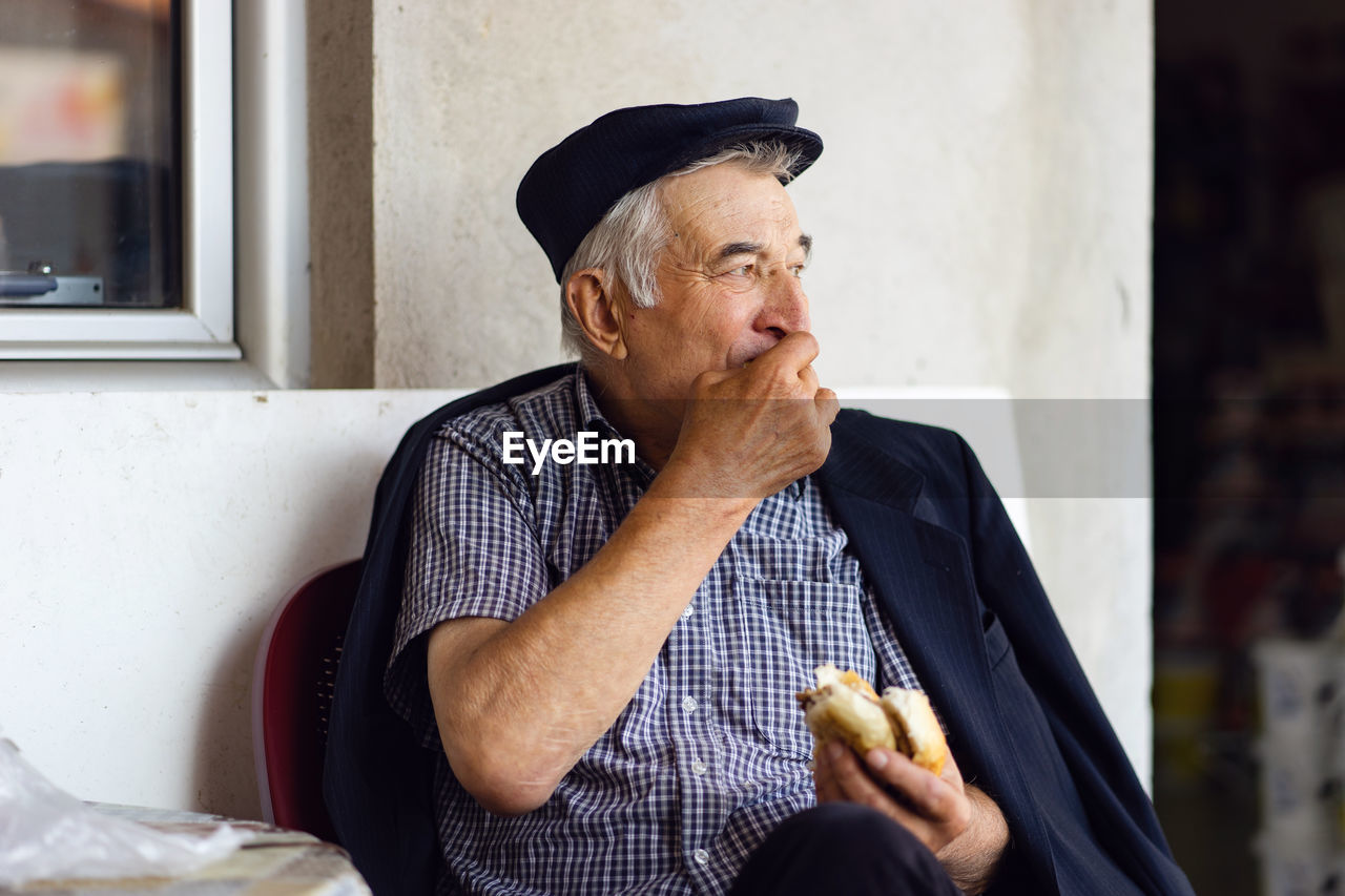 Senior man eating burger while sitting against wall