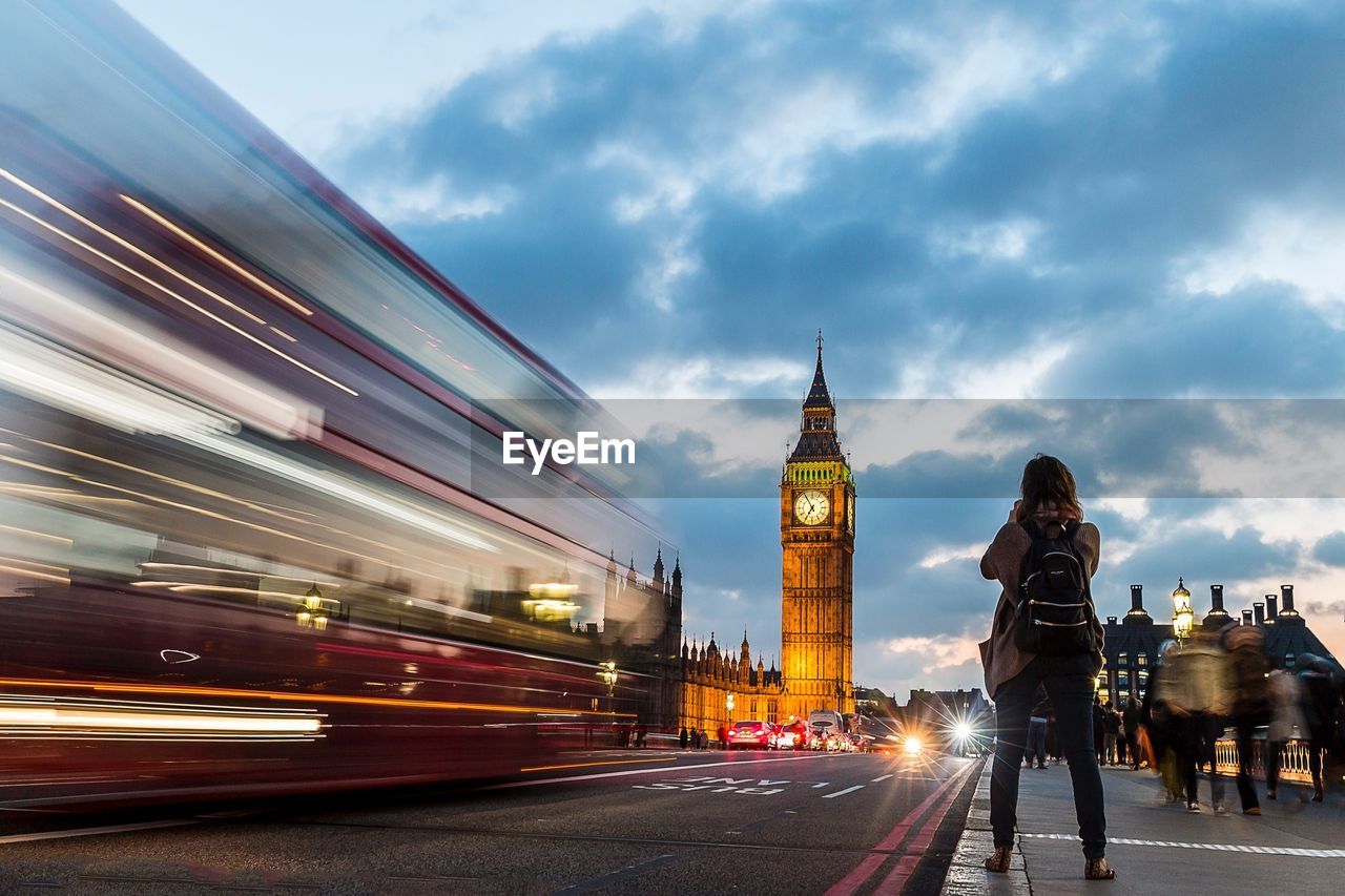 Rear view of woman standing on sidewalk by bus and big ben against cloudy sky during sunset