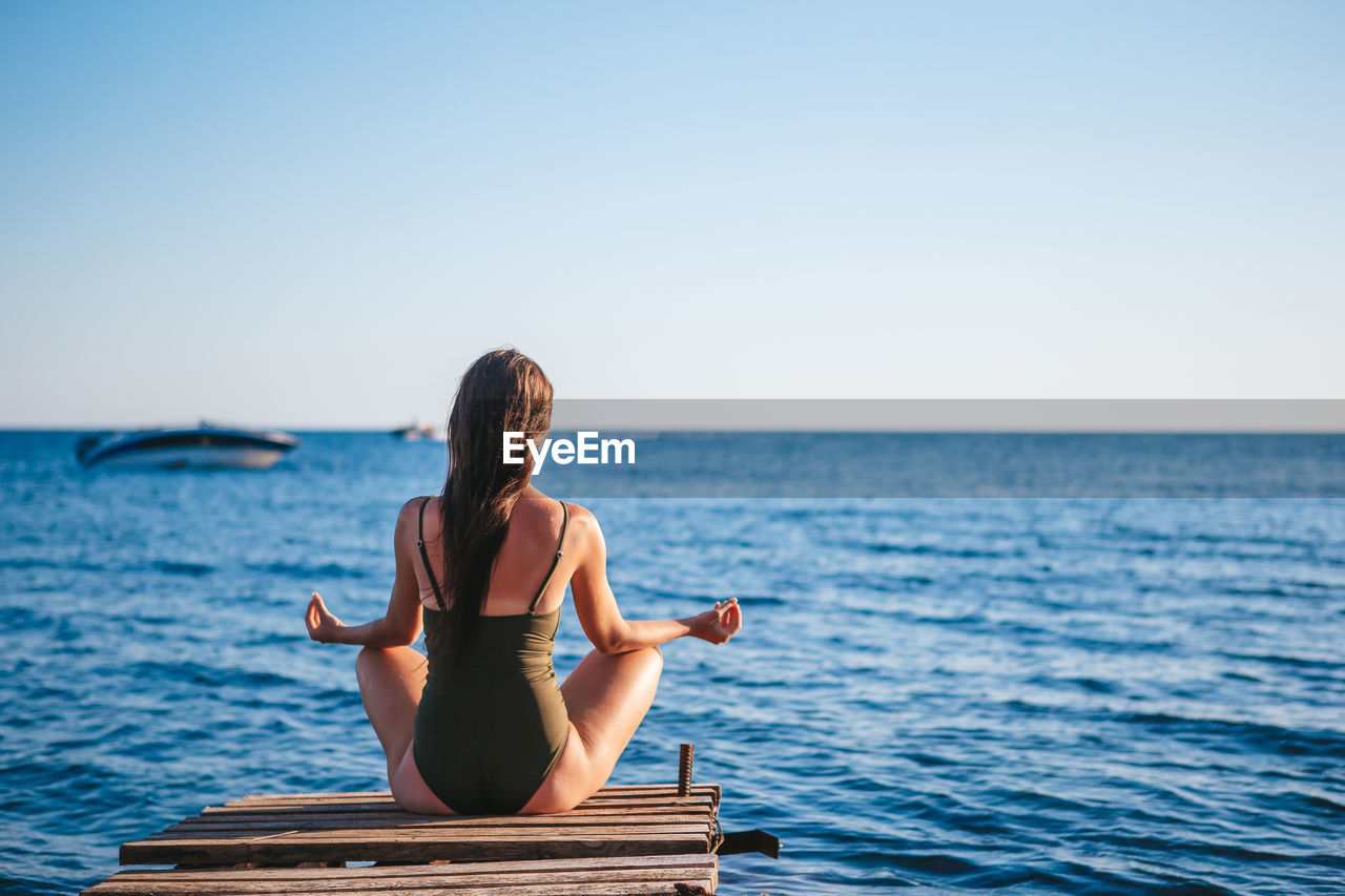 WOMAN SITTING AT SEA AGAINST CLEAR SKY