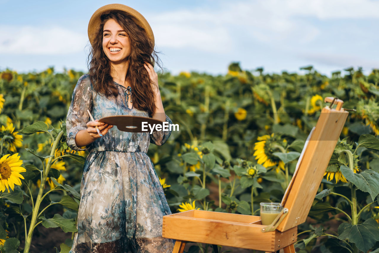 A young woman with curly hair and wearing a hat is painting in nature. 