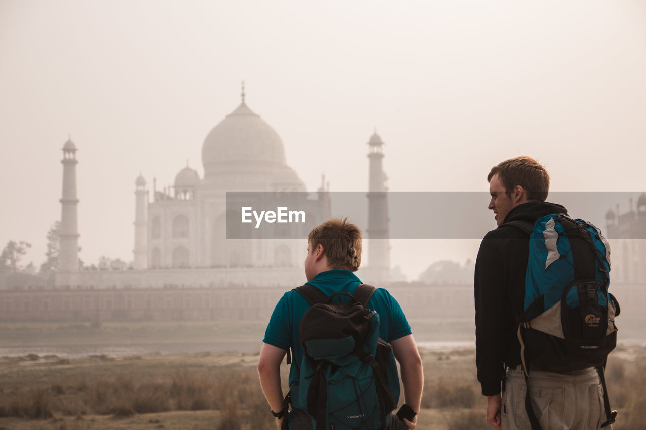 Rear view of tourists carrying backpacks while standing in front of taj mahal during foggy weather