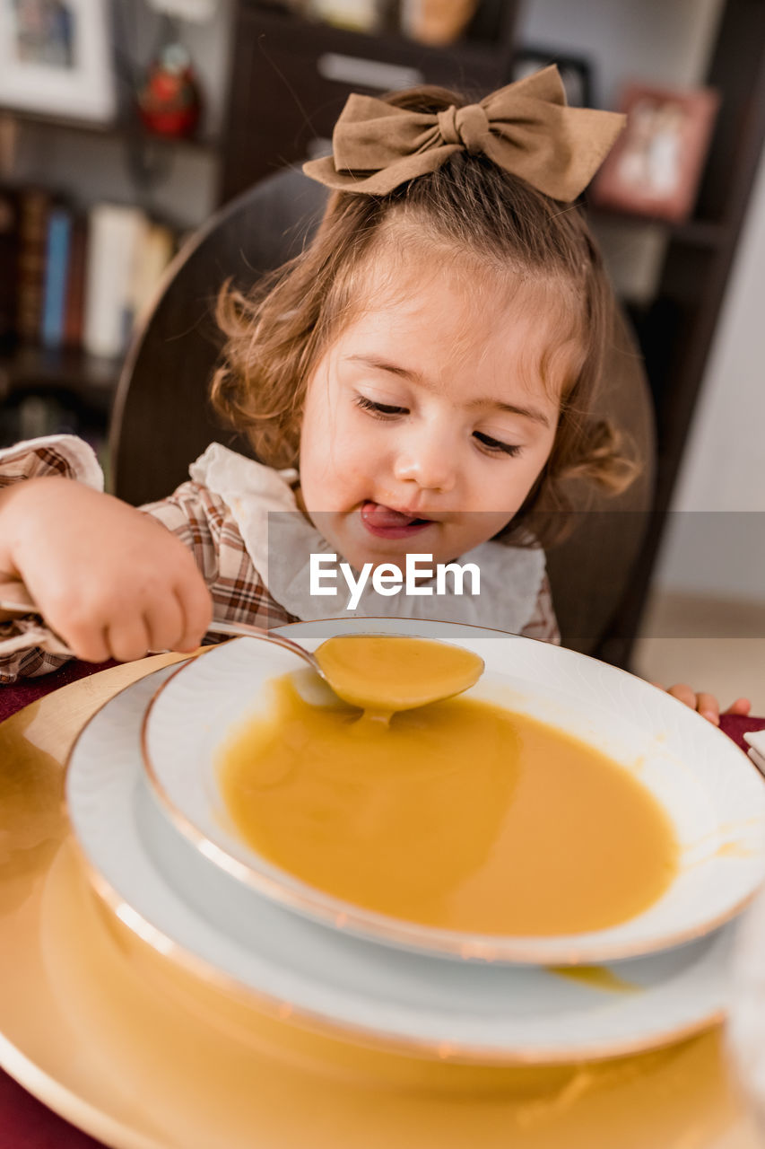 Charming child with bow on brown hair and spoon against plate of squash puree soup in house