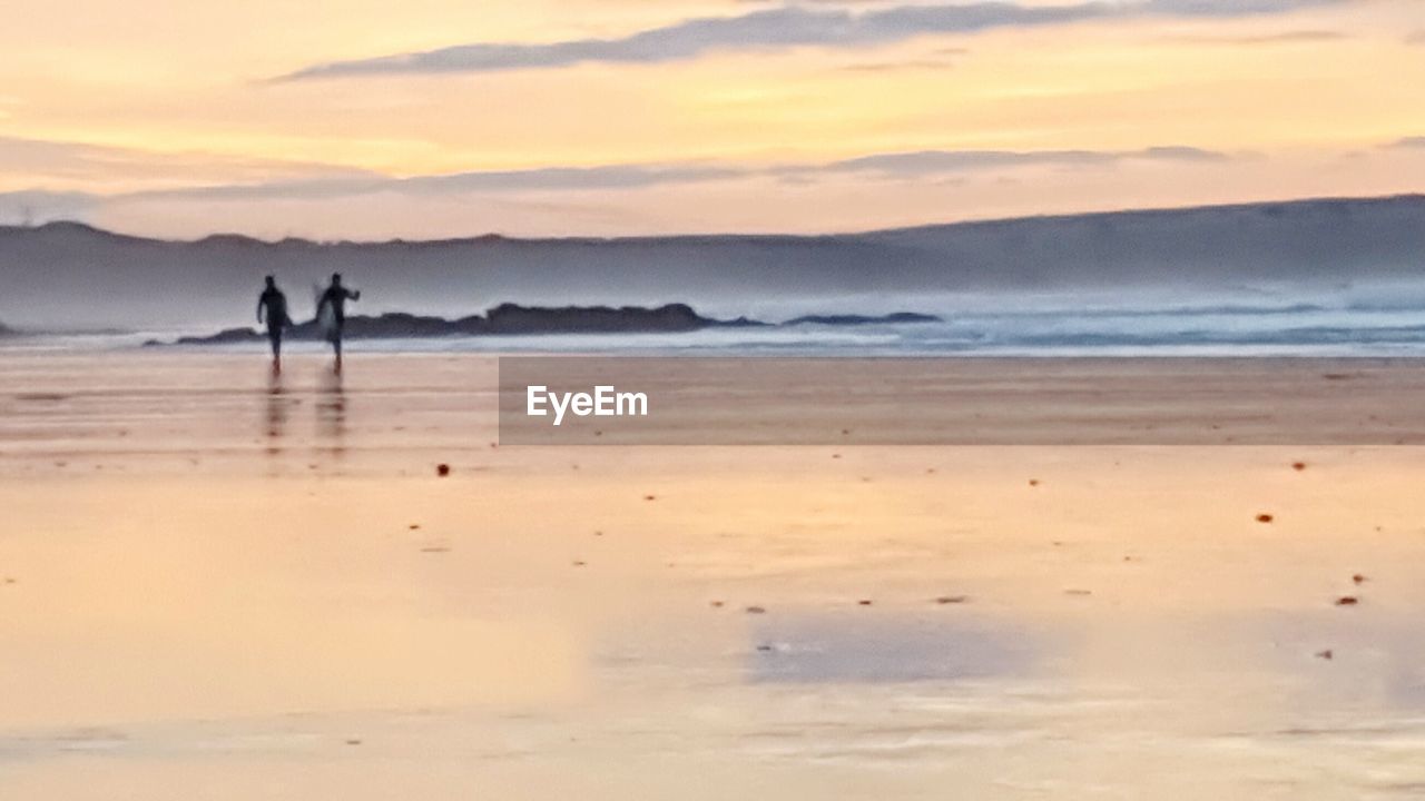 SILHOUETTE MAN WALKING ON BEACH AGAINST SKY AT SUNSET