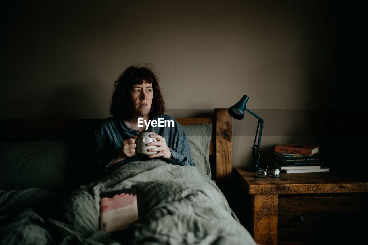 Woman holding coffee cup resting on bed at home