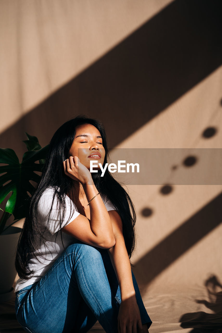 Attractive young hispanic woman wearing white cloth touching face with eyes closed in sun shadow