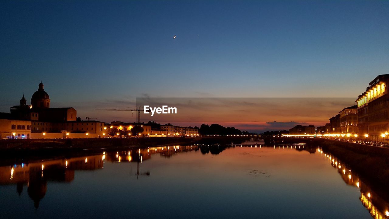 Illuminated buildings by river against sky at night