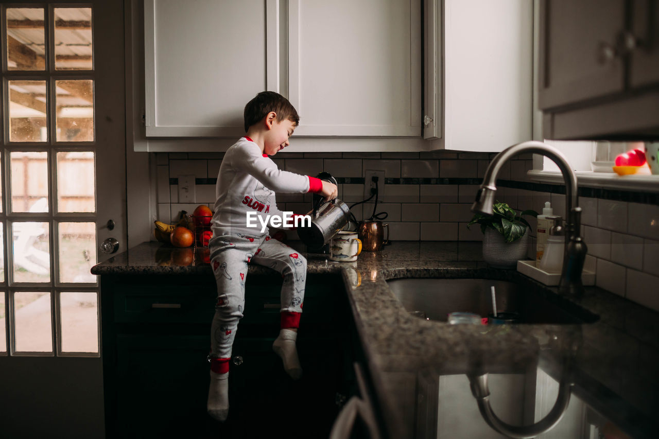 Young boy pouring flothed milk into cup of coffee on kitchen counter