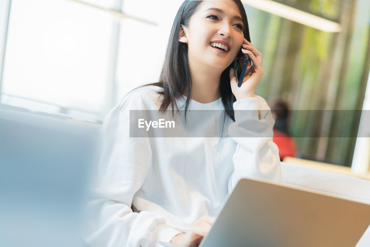 SMILING YOUNG WOMAN USING MOBILE PHONE IN LAPTOP