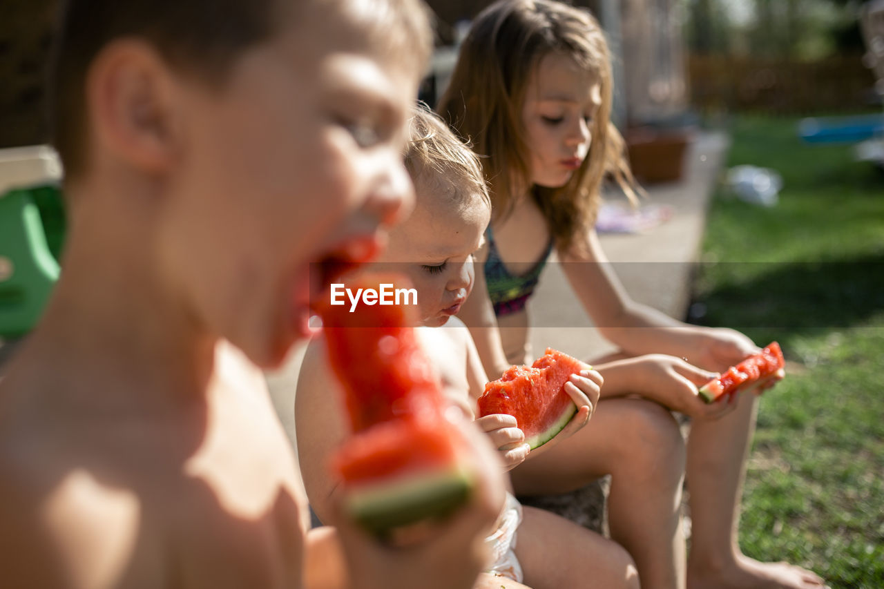 Siblings eating watermelons while sitting at yard