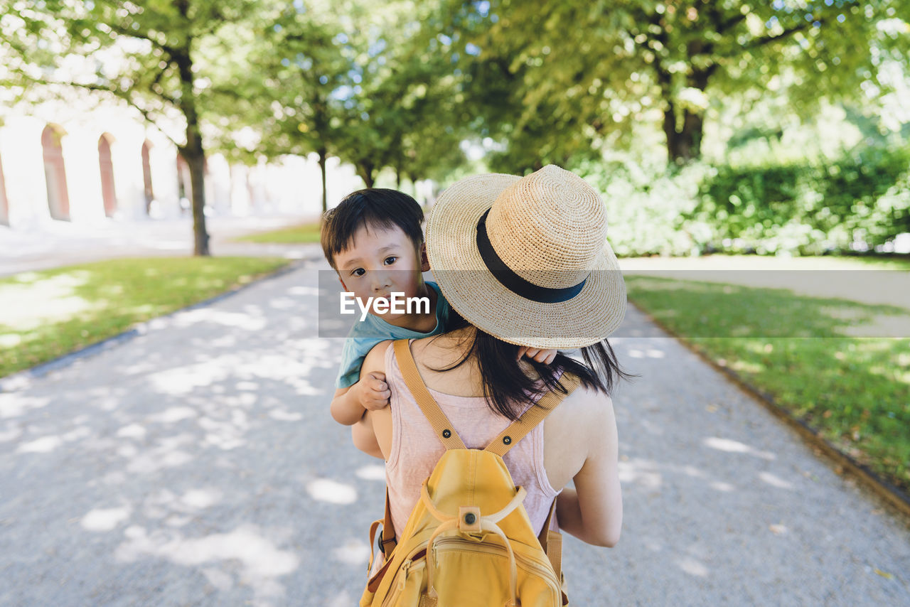 A boy looks back at the camera while his mother holding him walking toward a park.