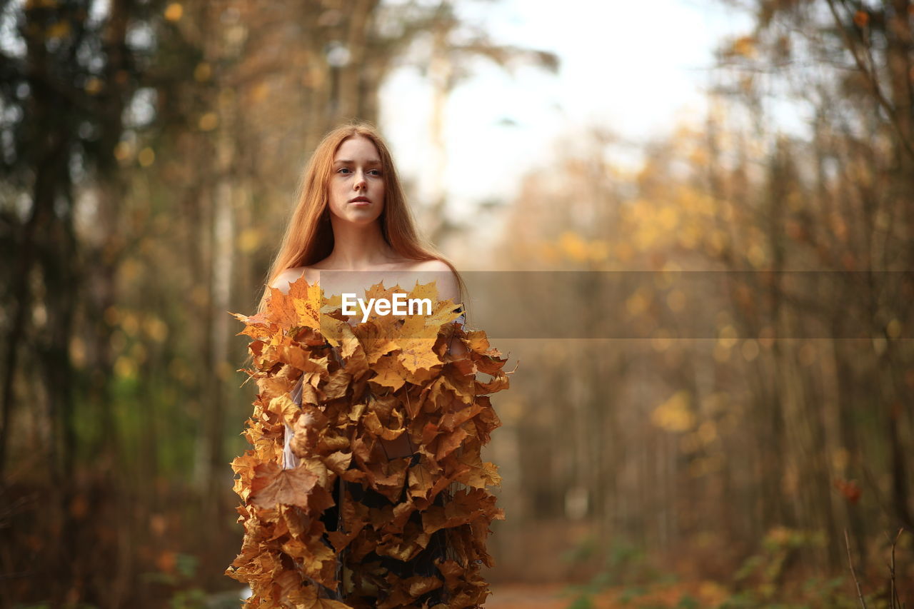 Thoughtful young woman covered with leaves standing at forest during autumn