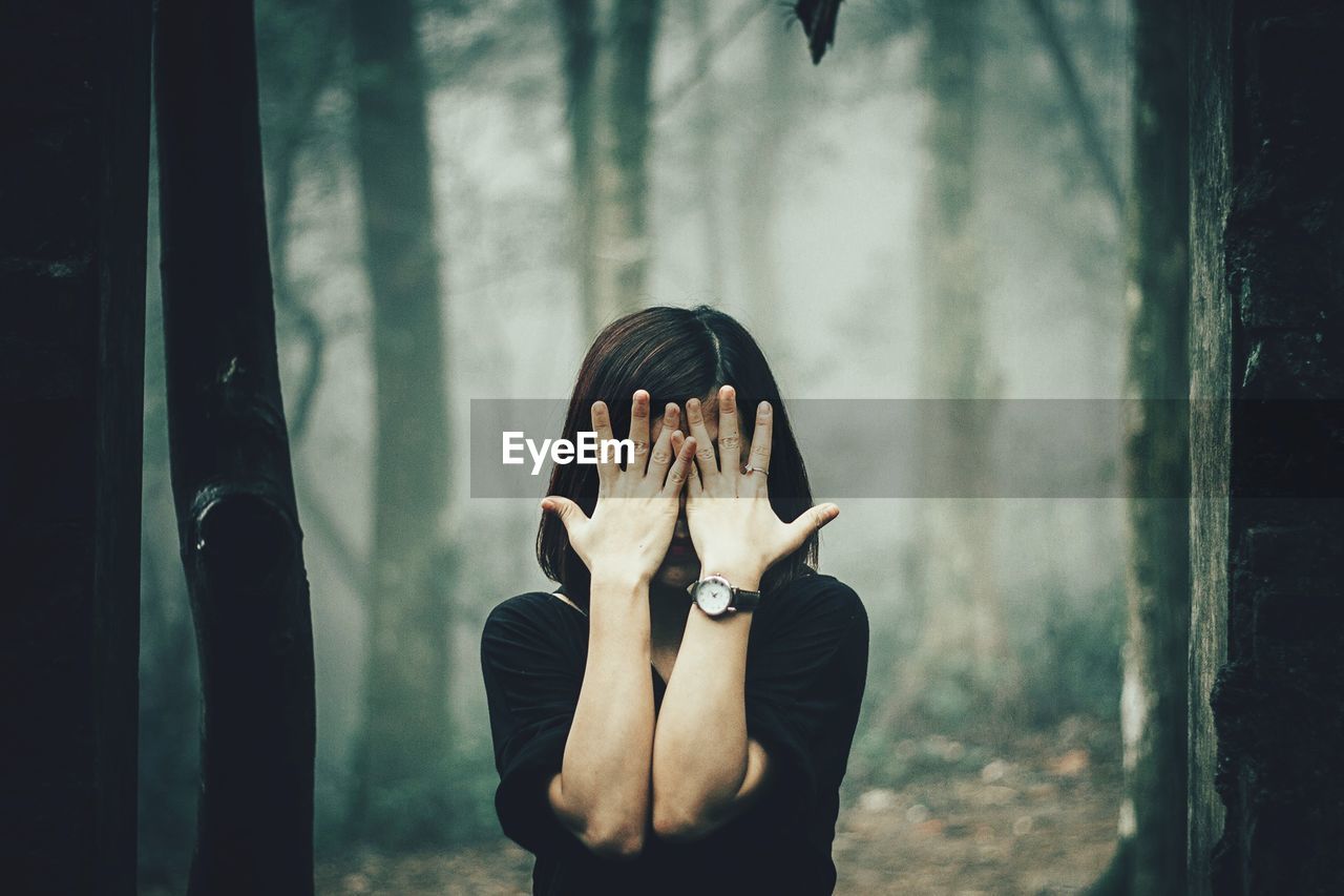 Woman covering face with hands while standing against trees