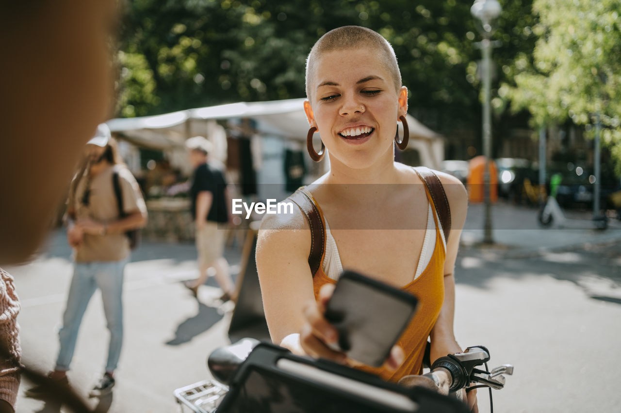 Smiling woman with shaved head paying via tap to pay while shopping at flea market