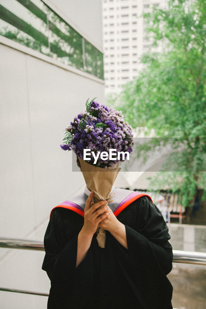 Person covering face with bouquet while wearing graduation gown against buildings in city