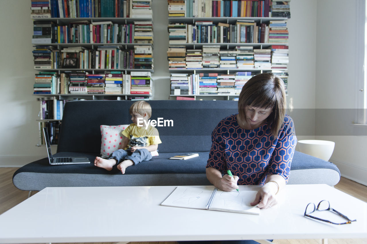 Mother writing while son looking laptop computer on sofa at home