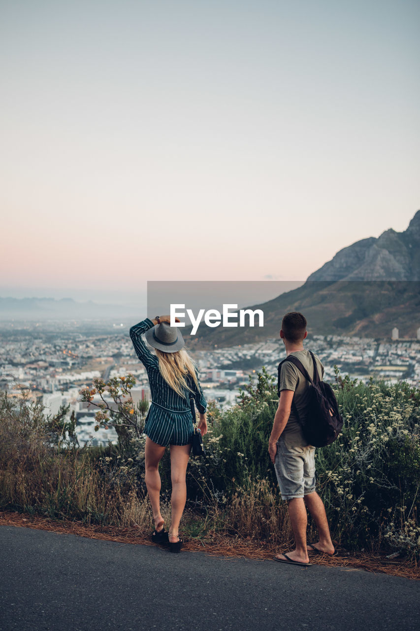 Rear view of friends standing on road by cityscape against clear sky during sunset