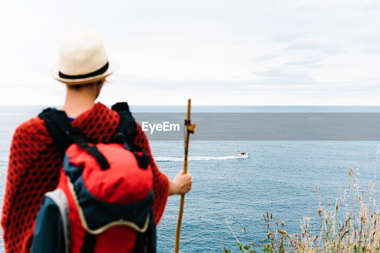 Back view of anonymous female hiker with backpack and stick standing on hill and admiring rippling sea with floating boat while resting during camino de santiago route in spain