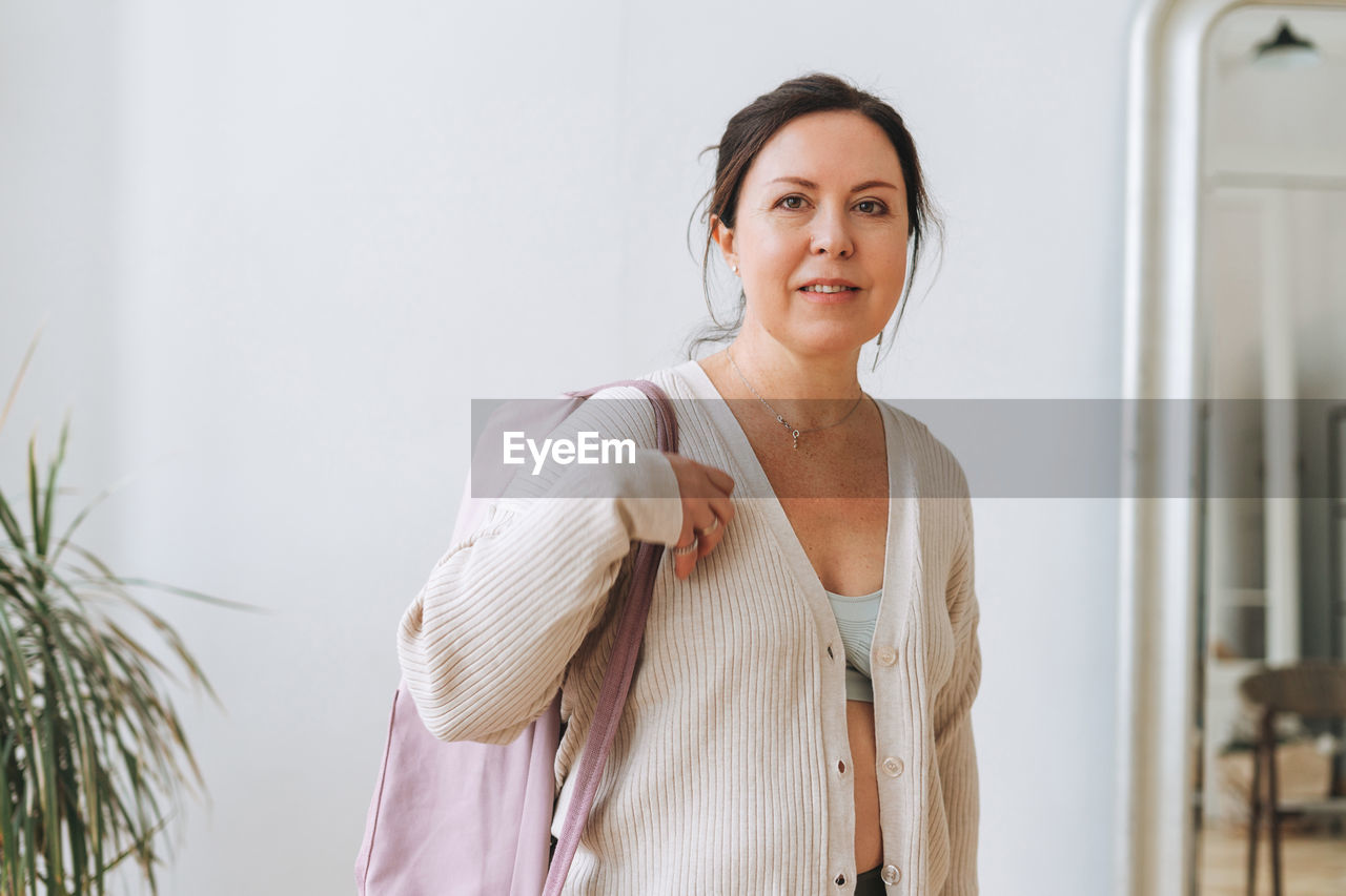 Attractive middle aged brunette woman in sportswear with yoga mat in light studio