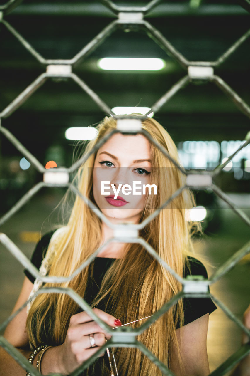 Portrait of young woman standing at parking garage seen through fence