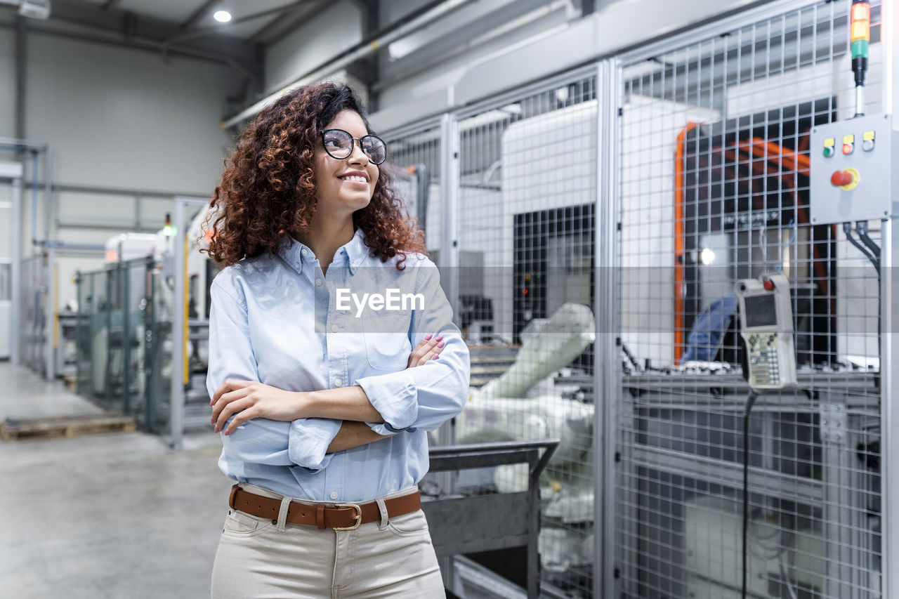 Happy young engineer with arms crossed standing in factory