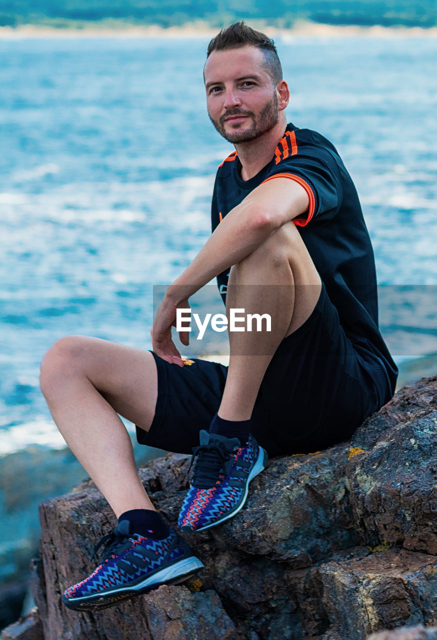 PORTRAIT OF YOUNG MAN SITTING ON ROCK AT SEA