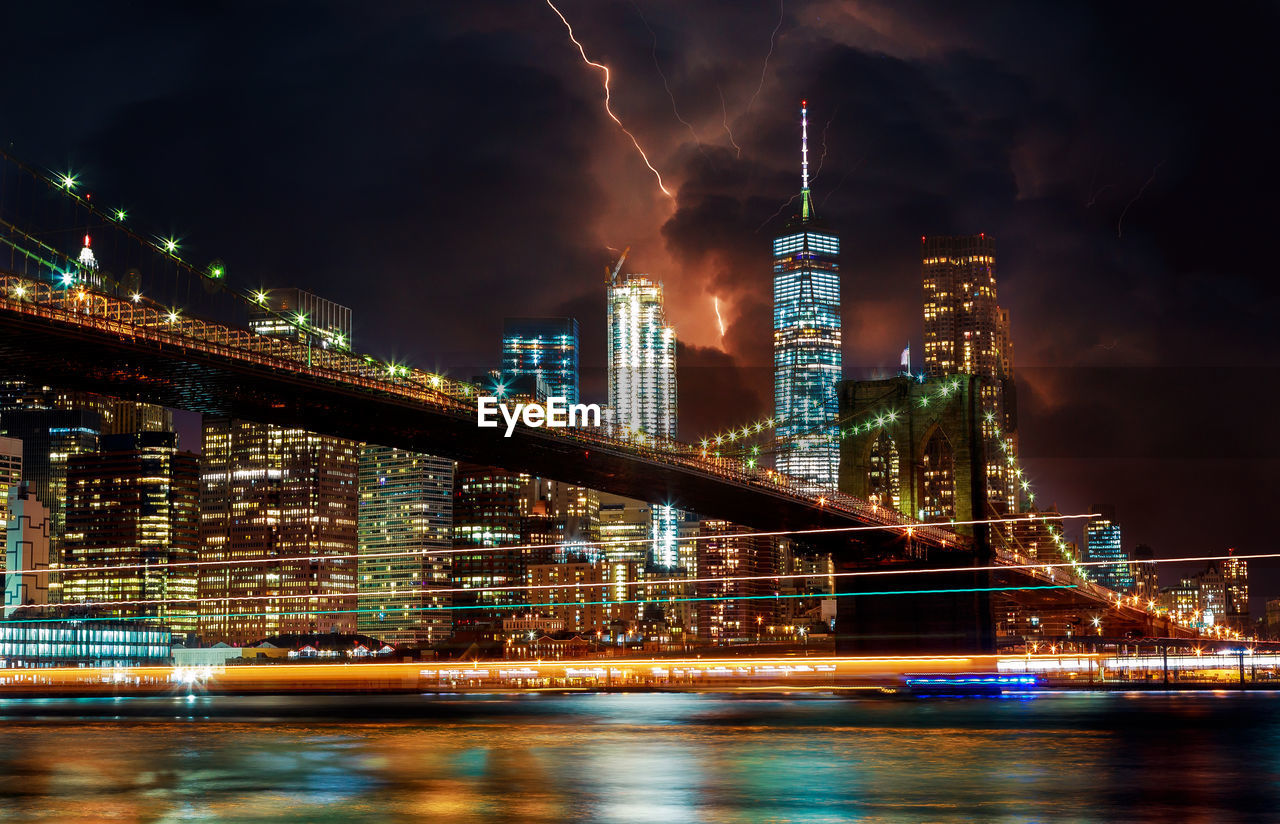 Illuminated bridge over river by buildings against sky at night