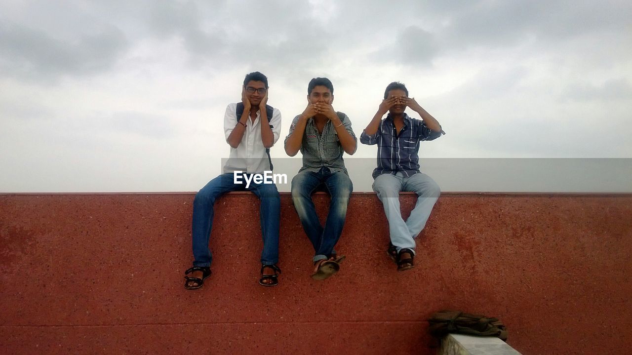 Full length of friends gesturing while sitting on retaining wall against sky