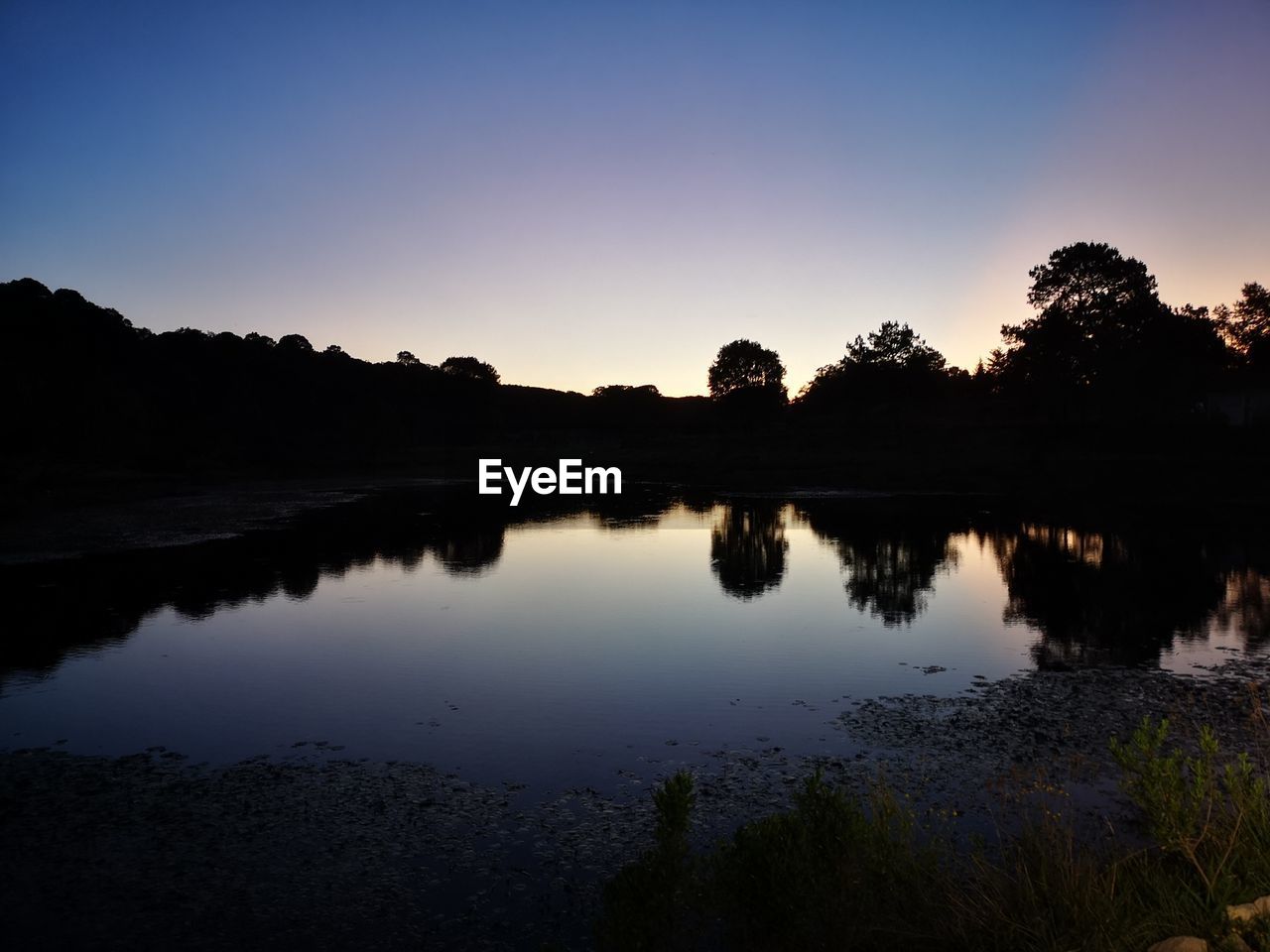 REFLECTION OF SILHOUETTE TREES IN LAKE AGAINST SKY DURING SUNSET