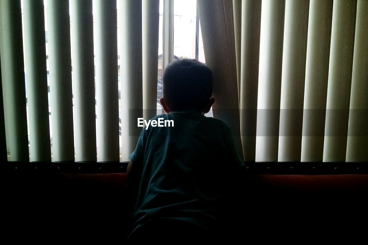 Rear view of boy looking through blinds window at home