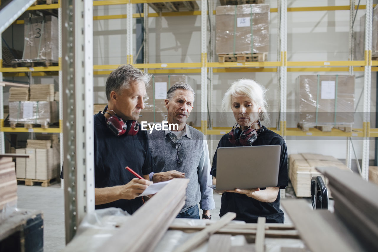 Female worker showing laptop to colleagues while standing by rack in industry