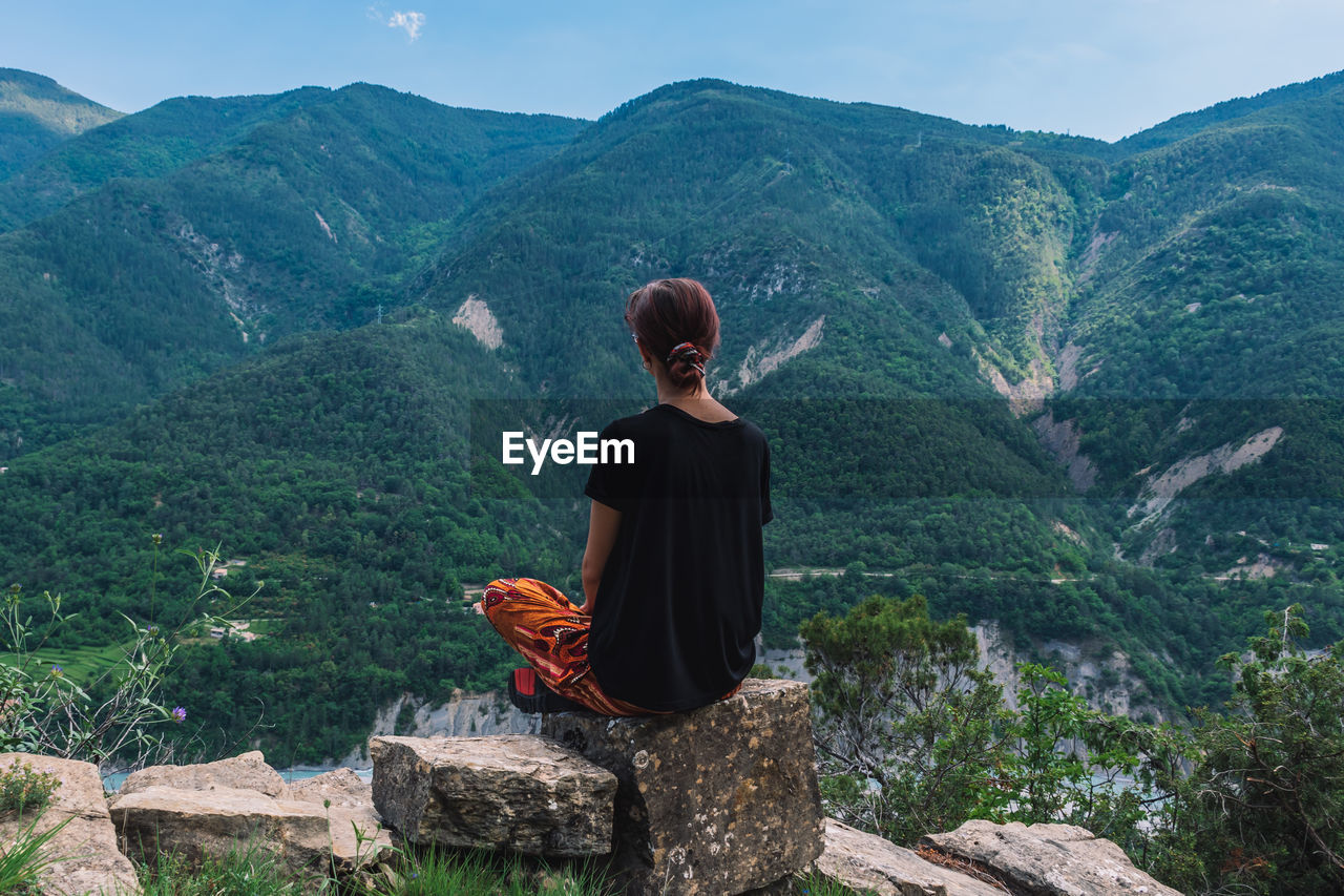 REAR VIEW OF MAN SITTING ON ROCK AGAINST MOUNTAINS