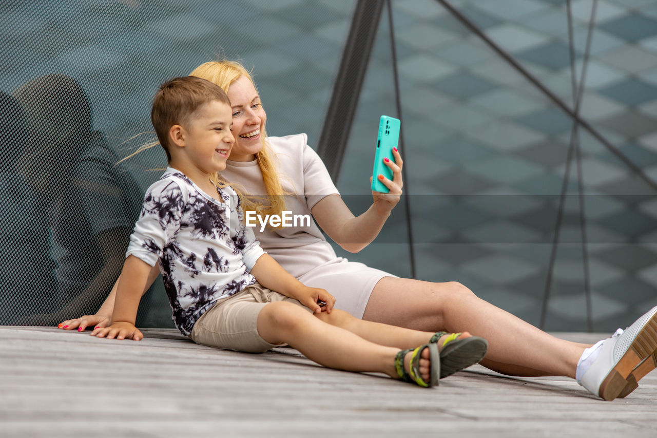 Happy blond woman and little boy sitting on terrace and making selfie on smartphone. mother and son