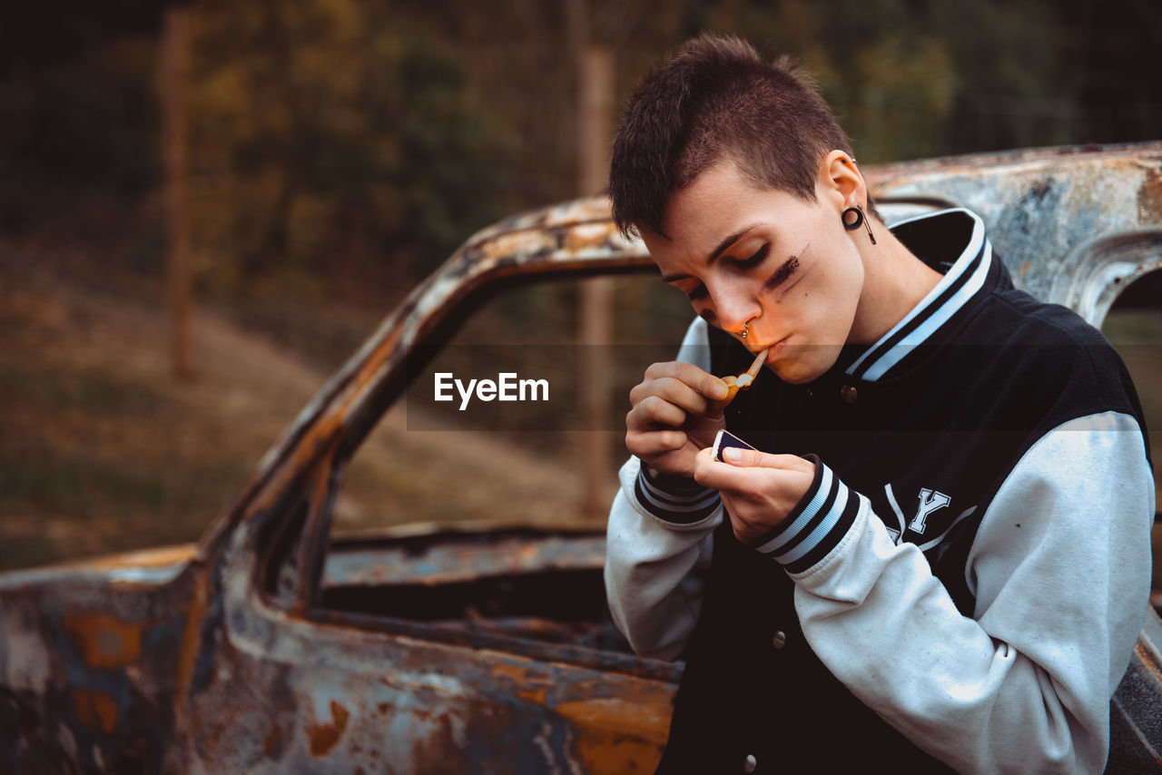 Young female with short hair and painted face lighting cigarette with match while standing near old rusty car in countryside and smoking