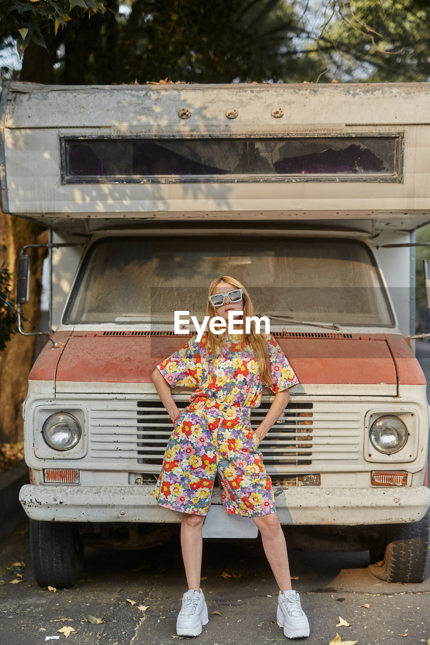 Confident female in trendy summer overall standing near old abandoned caravan and looking at camera