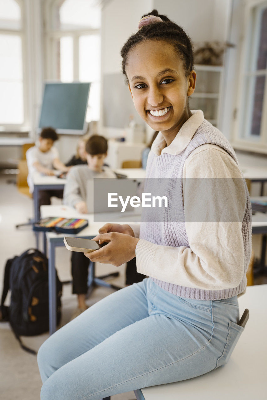 Portrait of happy schoolgirl holding smart phone while sitting at desk in classroom