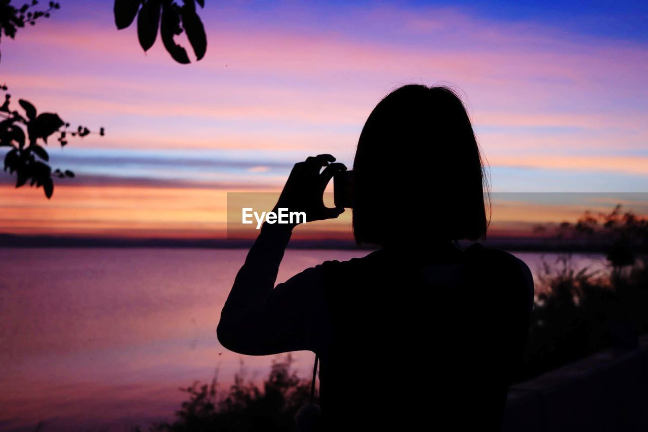 Silhouette woman photographing sea with smart phone against sky during sunset