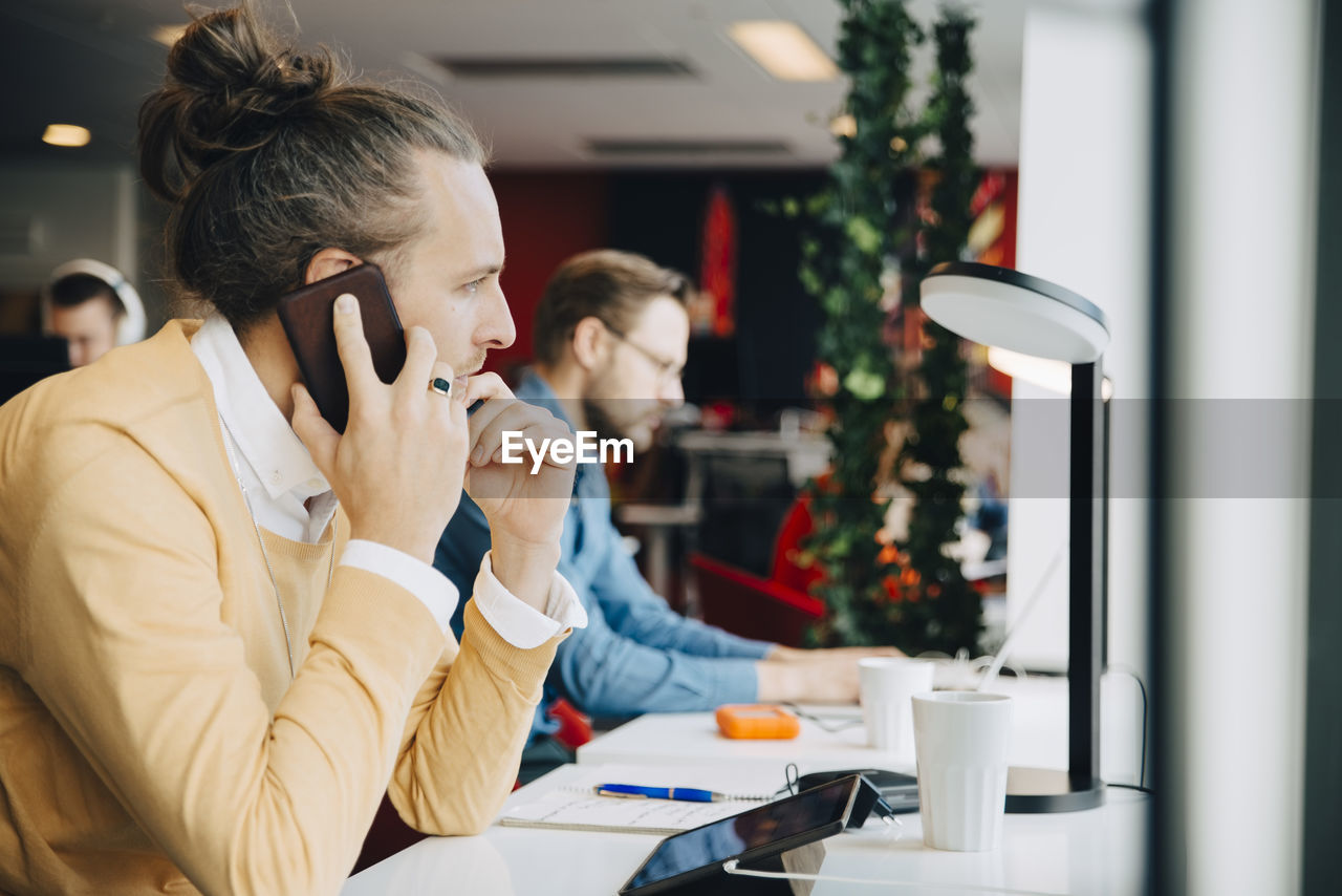 Side view of mid adult businessman talking on smart phone while sitting at desk in office