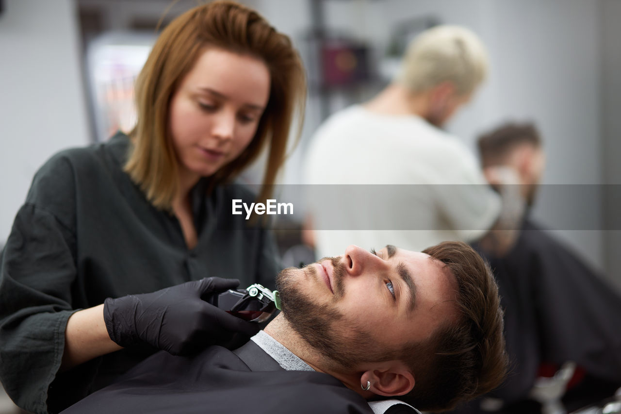 Portrait of young man who sitting in barbershop and woman hairdresser cut hair