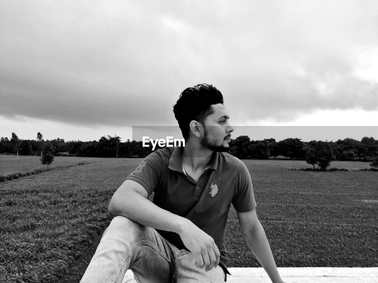YOUNG MAN LOOKING AWAY WHILE SITTING ON LAND AGAINST SKY