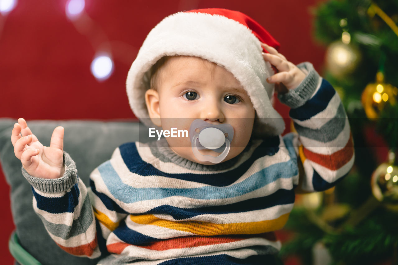 Portrait of cute baby boy with pacifier in mouth wearing santa hat