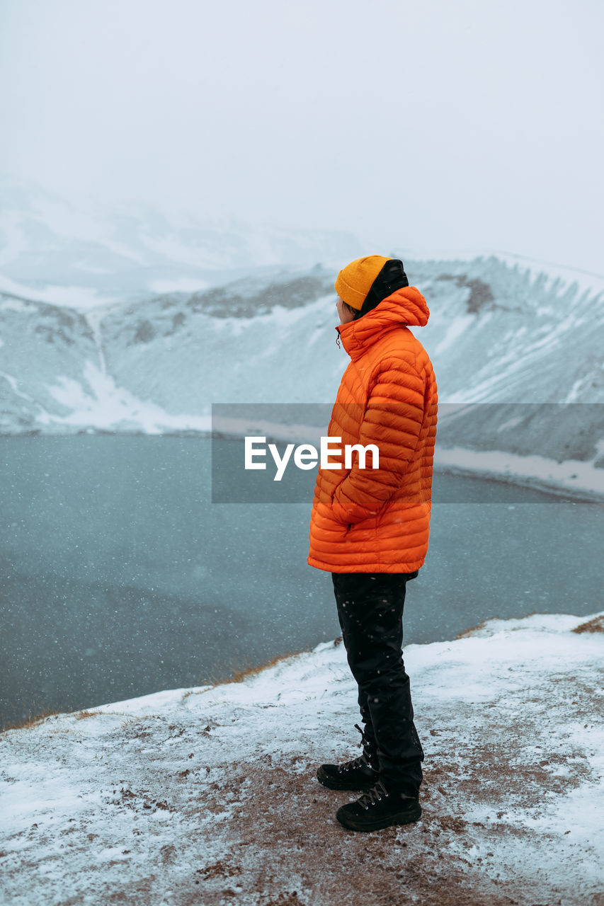 Side view of young tourist on peak of mountain in snow looking at water in valley