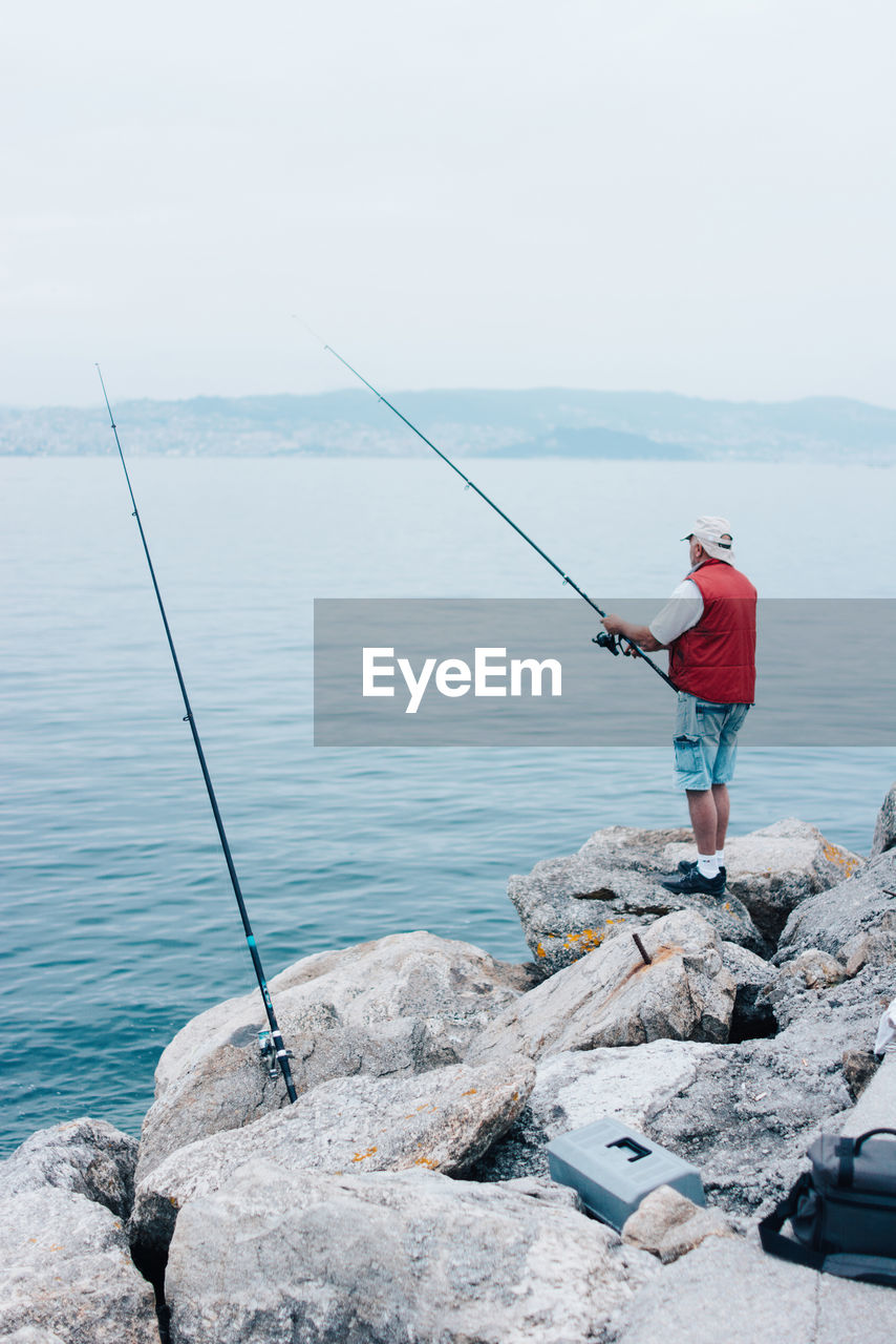 Side view of active man in red waistcoat with fishing rod standing on stony shoe and fishing in bright day