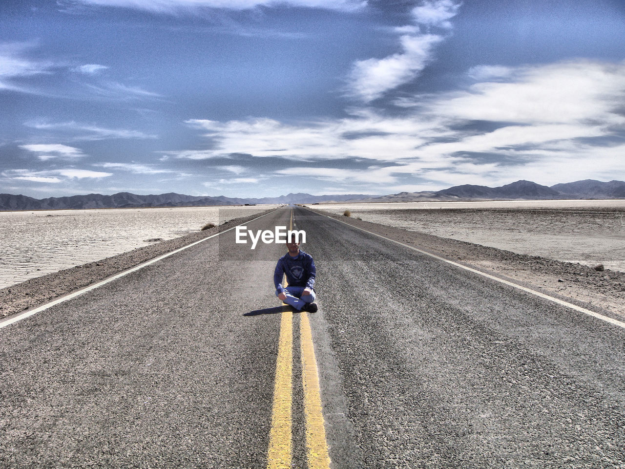 Man sitting on road leading towards mountains against blue sky