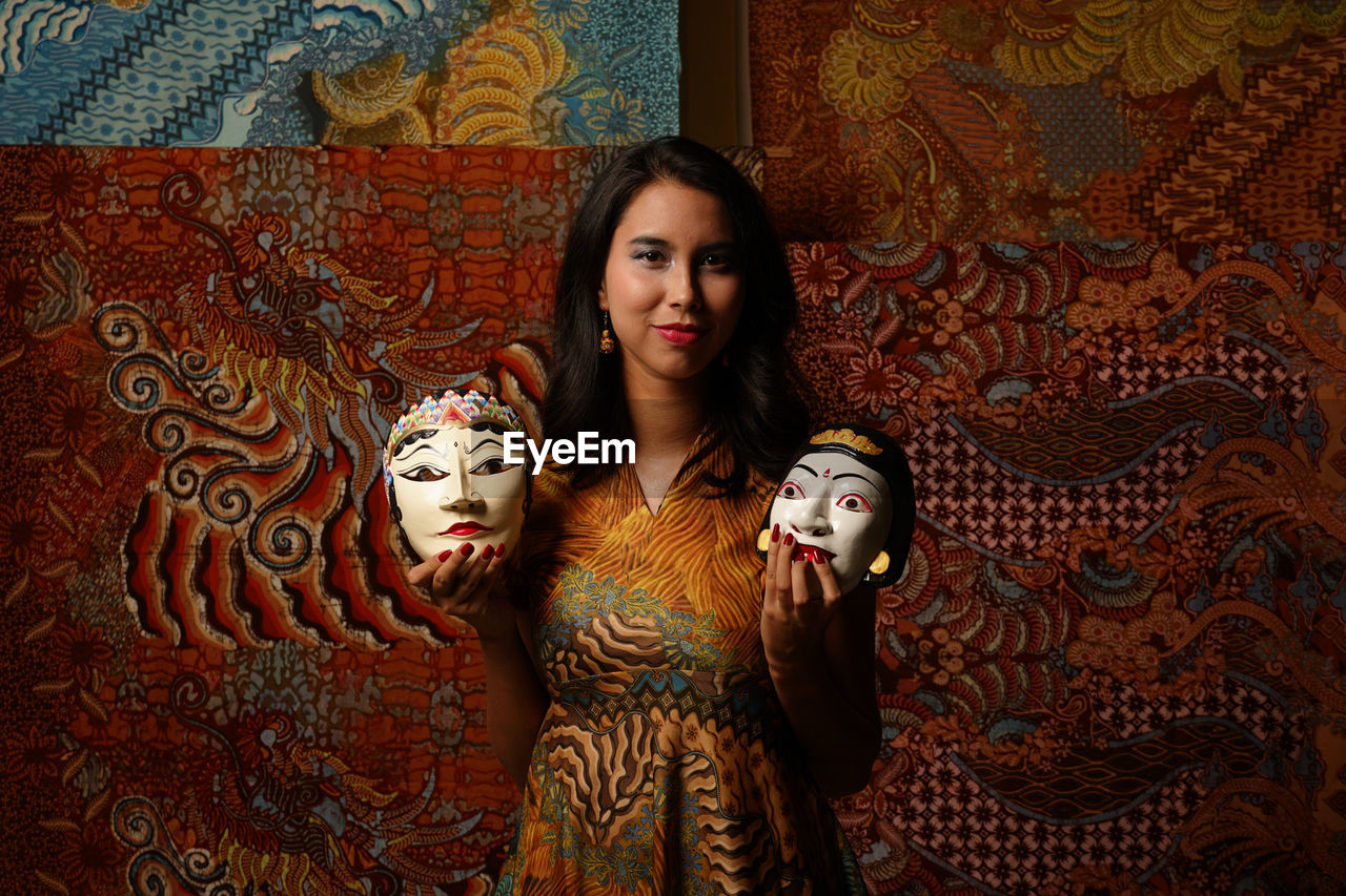 Portrait of smiling young woman holding mask against wall