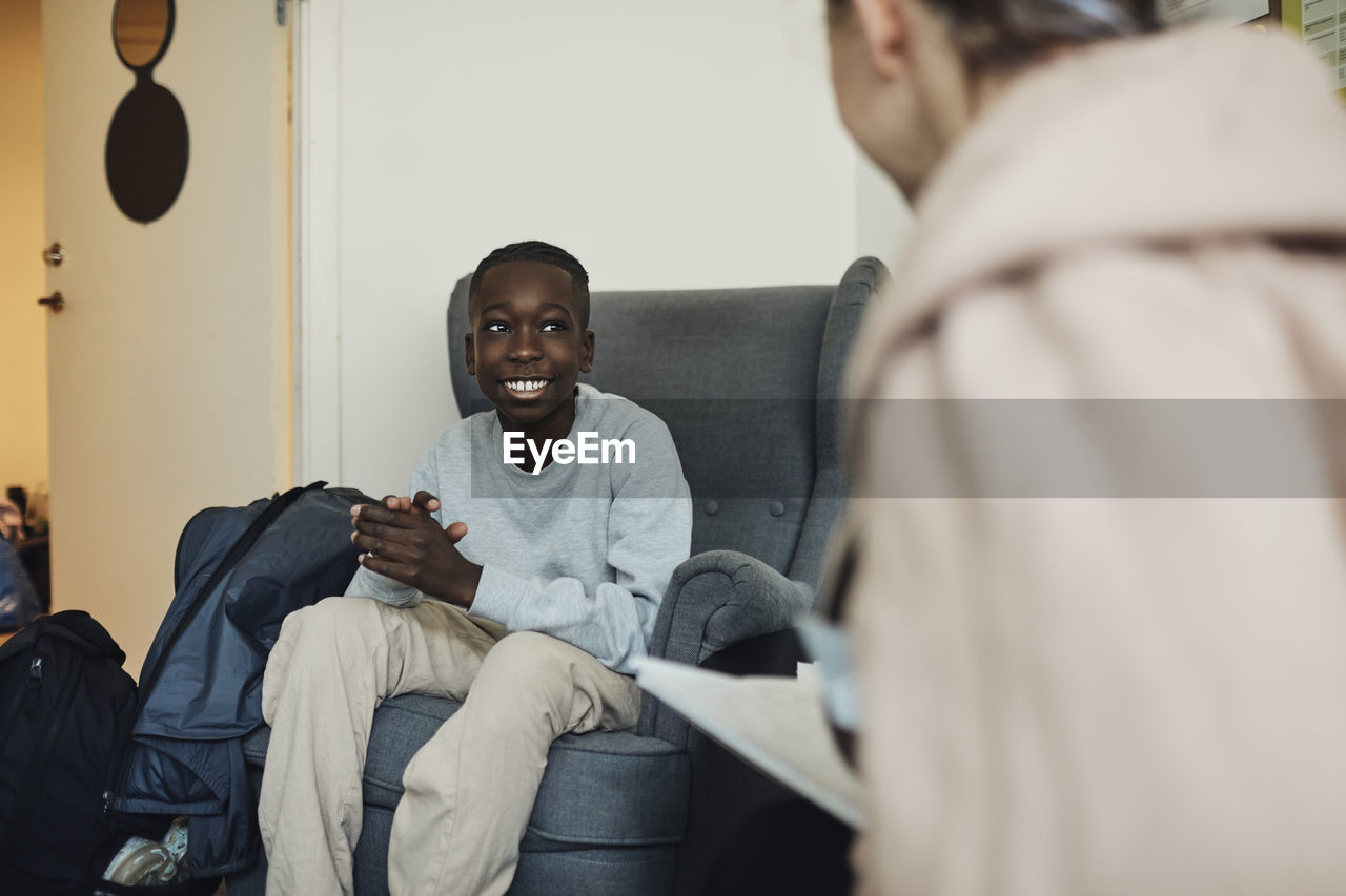 Smiling teenage boy talking with mental health professional while sitting on chair in school office