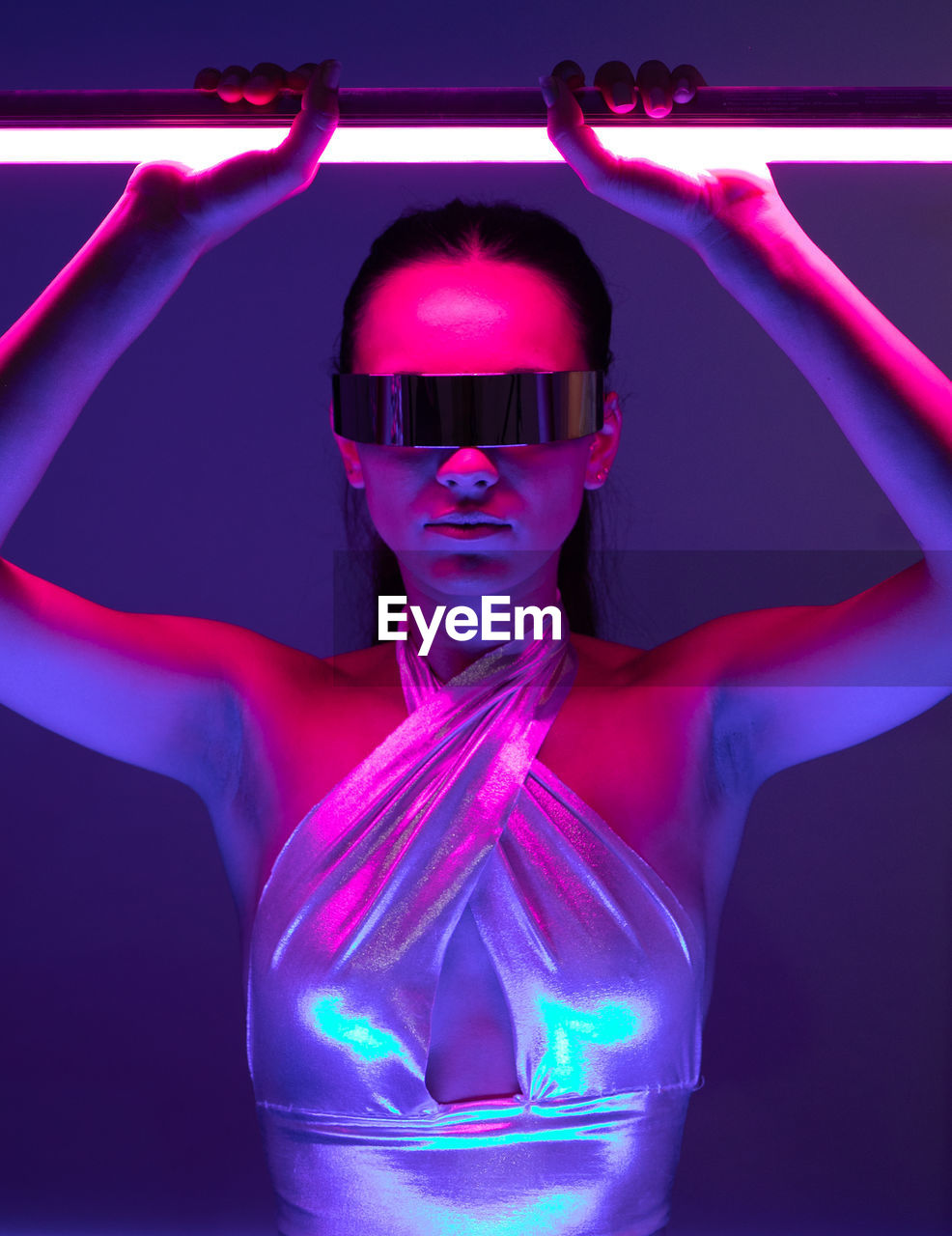 Self assured young female model with long hair in futuristic outfit and vr goggles standing in dark room with neon sword in hands