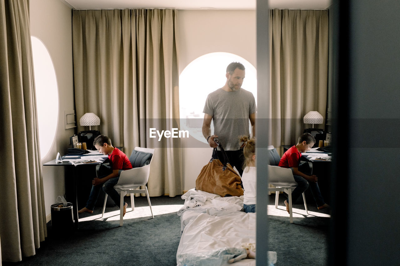 Man holding bag by bed while boy sitting at table in hotel