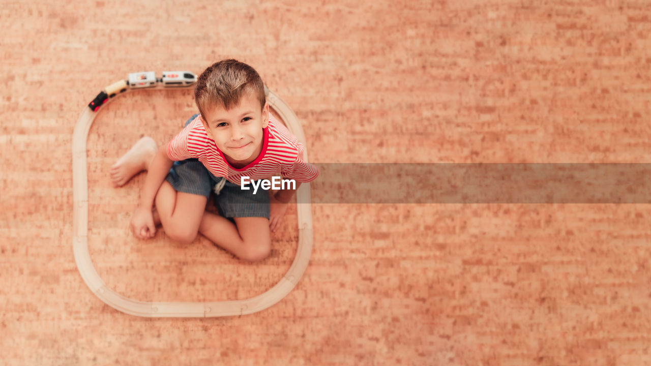 Cute little boy sitting on floor, around him is wooden railway with a train. child looking at camera