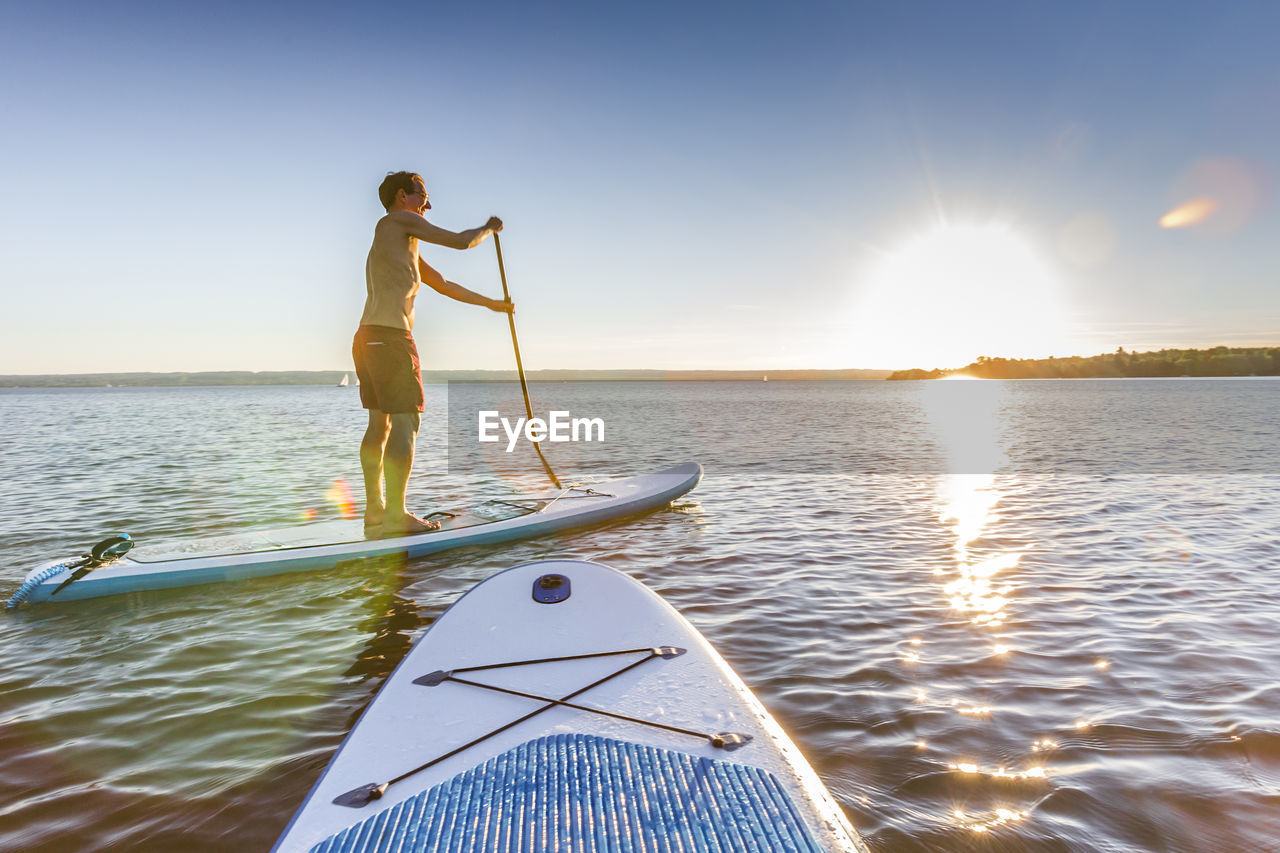 Side view of shirtless man paddleboarding on river against sky during sunset